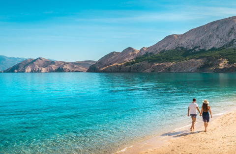 Una coppia passeggia mano nella mano sulla spiaggia di Baška, Croazia, con mare azzurro e montagne.