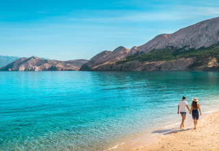 Una coppia passeggia mano nella mano sulla spiaggia di Baška, Croazia, con mare azzurro e montagne.