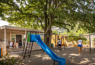 Playground with slide and children playing under trees at Campsite «Baška» holiday park in Primorje-Gorski Kotar, Croatia.
