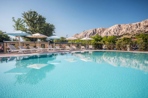 Piscina all'aperto con lettini e ombrelloni al campeggio Baška in Croazia, montagne visibili sullo sfondo.