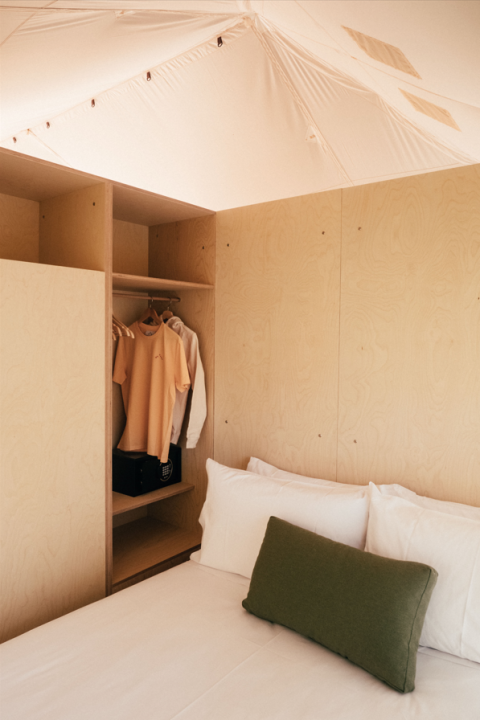 Interieur van een safaritent met houten panelen, bed met groen kussen en open kledingkast met kleding.