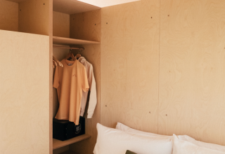 Interieur van een safaritent met houten panelen, een bed met groen kussen en open garderobe met kleren.