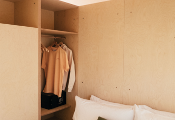 Interno di una tenda safari con pannelli in legno, letto con cuscino verde e guardaroba aperto con vestiti.