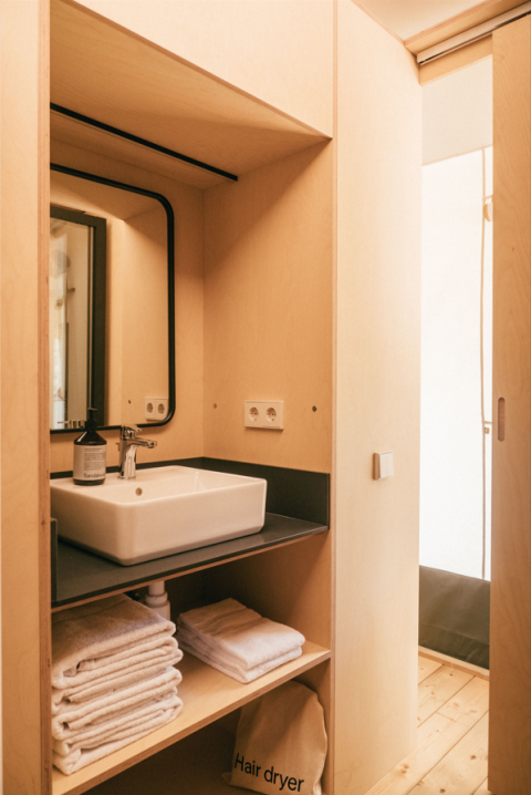Badkamer in safaritent met wastafel, spiegel, stapels handdoeken en haardrogerzak op houten planken.