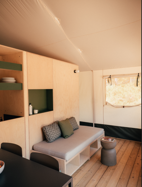 Interior of a safari tent featuring light wooden furniture, a sofa, shelves, table, and a sunlit window.