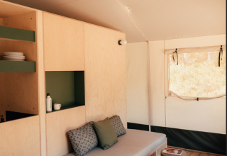 Interior of a safari tent featuring light wooden furniture, a sofa, shelves, table, and a sunlit window.