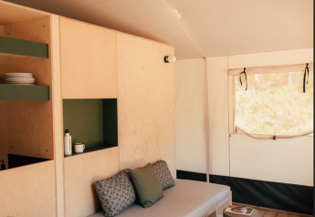 Interno di una tenda safari con mobili in legno chiaro, divano, scaffali, tavolo e finestra soleggiata.
