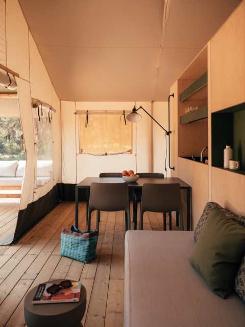 Interieur van een safaritent met houten vloer, eetruimte, comfortabele zetel en open kitchenette.