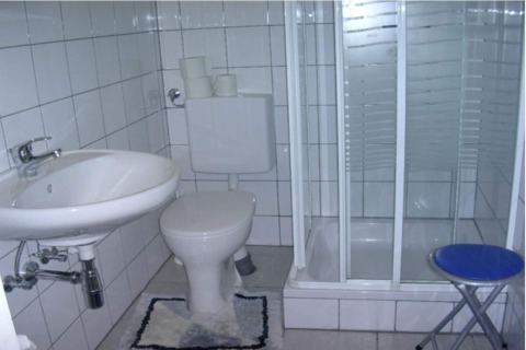 Et lille badeværelse i en hytte med håndvask, toilet, brusekabine, toiletpapir og en blå klapstol.