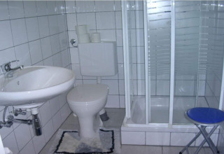 Et lille badeværelse i en hytte med håndvask, toilet, brusekabine, toiletpapir og en blå klapstol.