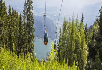 A cable car cabin glides over lush green trees, overlooking a scenic lake and distant mountains.