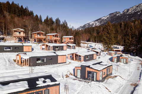 Glamping invernale con moderne cabine di legno immerse in un paesaggio montano innevato e alberi di pino.