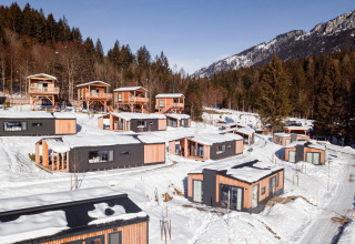 Glamping invernale con moderne cabine di legno immerse in un paesaggio montano innevato e alberi di pino.