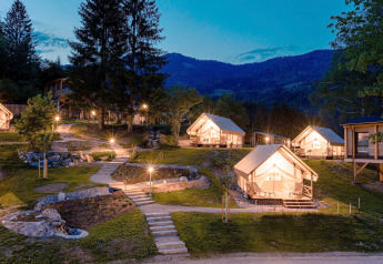 Glampingaccommodatie met verlichte luxe tenten in de avond, omgeven door natuur en bergen op de achtergrond.
