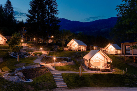 Glamping di notte con tende di lusso illuminate, sentieri in pietra e montagne visibili sullo sfondo.