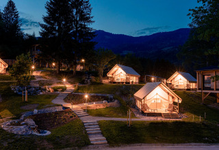 Glamping di notte con tende di lusso illuminate, sentieri in pietra e montagne visibili sullo sfondo.