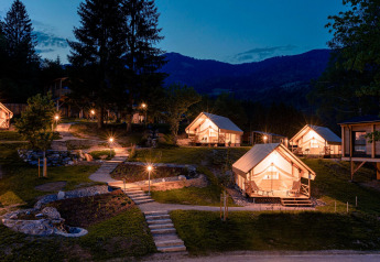 Glampingaccommodatie met verlichte luxe tenten in de avond, omgeven door natuur en bergen op de achtergrond.