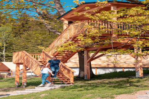 Una coppia scende dalla casa sull’albero in legno in un glamping immerso nella natura e tra alberi verdi.
