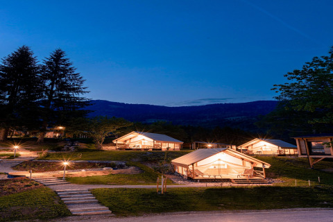 Tende glamping illuminate di notte su terrazze erbose in montagna, con sentieri e alberi intorno.