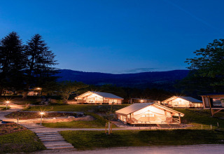 Tende glamping illuminate di notte su terrazze erbose in montagna, con sentieri e alberi intorno.