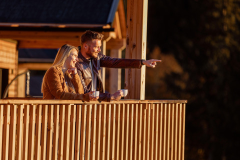 Una coppia si gode il caffè del mattino sulla terrazza del loro alloggio glamping, sorridendo al panorama.
