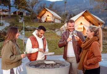 Vier vrienden drinken wijn en grillen bij een vuurplaats voor luxe glampingtenten in de natuur.