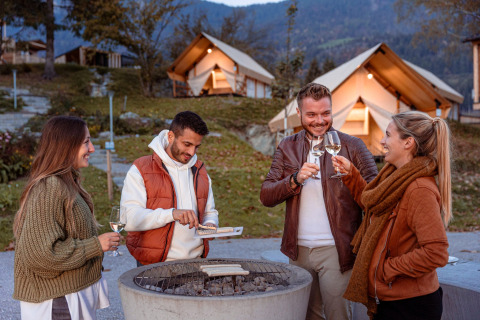Quattro amici si godono vino e cibo attorno al fuoco davanti alle tende di un glamping panoramico.