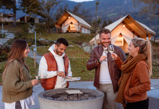 Quattro amici si godono vino e cibo attorno al fuoco davanti alle tende di un glamping panoramico.