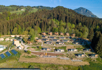 Luchtfoto van een glampinglocatie met houten huisjes en tenten, omgeven door bos en bergen.