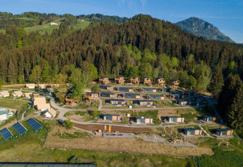 Luchtfoto van een glampinglocatie met houten huisjes en tenten, omgeven door bos en bergen.