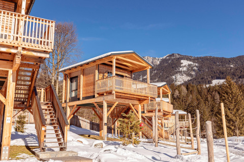 Træchalet på pæle i sneklædte bjerge med blå himmel, balkon og trapper, omgivet af fyrretræer.