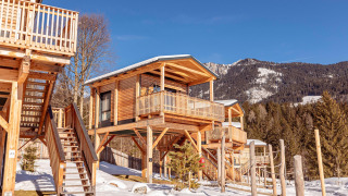Houten chalet op palen in een besneeuwd berglandschap, met een balkon, trap en dennenbomen.