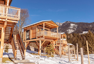 Træchalet på pæle i sneklædte bjerge med blå himmel, balkon og trapper, omgivet af fyrretræer.