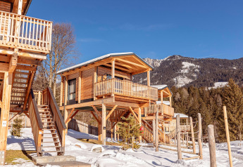 Chalet in legno su palafitte tra le montagne innevate, con balcone, scale e circondato da pini.