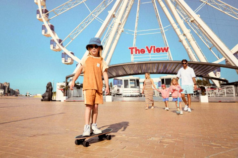 Familie voor het reuzenrad ‘The View’, kind skate op het plein terwijl anderen wandelen in de zon.