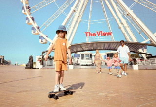 Famiglia davanti alla ruota panoramica 'The View', bambino sullo skateboard su una piazza soleggiata.