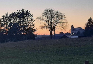 Foto di un alloggio glamping al tramonto con prati, alberi e una chiesa di paese sullo sfondo.