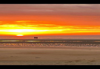Due persone ammirano un tramonto mozzafiato sulla spiaggia da una sistemazione glamping, cielo arancione acceso.