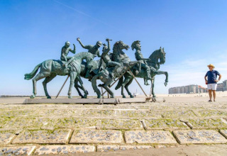 Ein Mann steht an einem Strand neben einer großen Reiterstatue, im Hintergrund Gebäude und blauer Himmel.