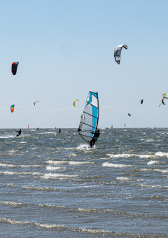 Windsurfistas y kitesurfistas en el mar, junto a un parque de vacaciones con glamping y cielo azul.