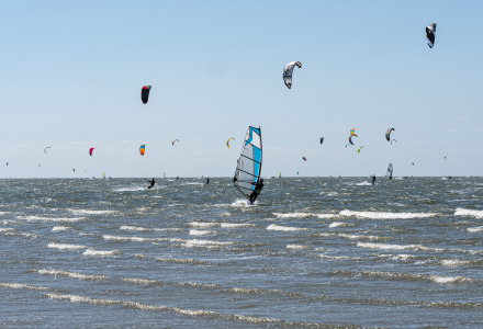 Windsurfistas y kitesurfistas en el mar, junto a un parque de vacaciones con glamping y cielo azul.