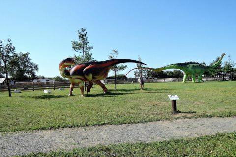Exhibición al aire libre con grandes estatuas de dinosaurios en césped, árboles y cielo azul de fondo.