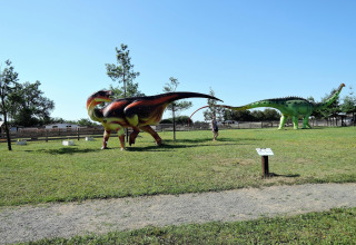 Exhibición al aire libre con grandes estatuas de dinosaurios en césped, árboles y cielo azul de fondo.
