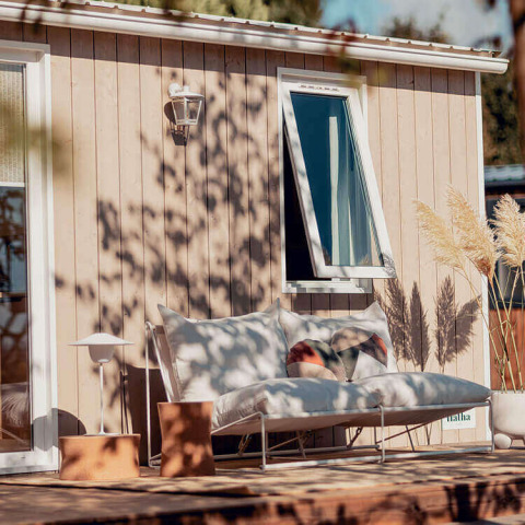 Gemütlicher Außenbereich eines Glamping-Zimmers mit Sofa, Kissen, Pflanzen und offenem Fenster.