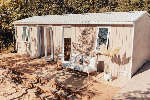 Buitenaanzicht van een glampingaccommodatie met houten terras, zitbank en planten, omgeven door groen.