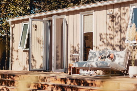 Zonnige glampingplek met houten terras, gezellige ligbedden en open deuren naar de natuur.