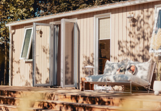 Zonnige glampingplek met houten terras, gezellige ligbedden en open deuren naar de natuur.