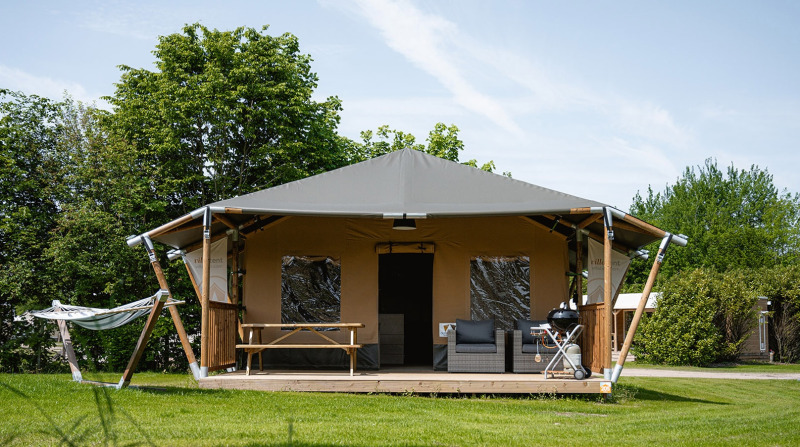 Tienda safari llamada Cottage con porche, hamaca, zona de estar y barbacoa en césped verde.