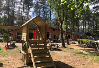 Alloggio glamping con area giochi, casetta di legno e posti a sedere all'aperto tra alberi e natura.