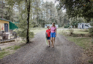 Une femme et un enfant marchent sur un chemin de gravier près d’un chalet avec des arbres et camping-cars autour.